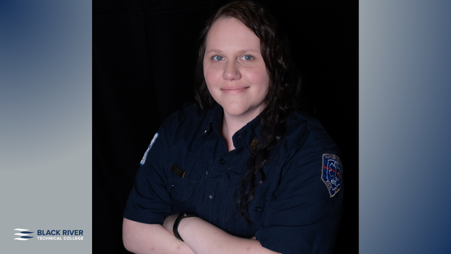 A woman with long dark curly hair, wearing a dark EMS uniform with a patch on the sleeve, poses with arms crossed against a black background, with Black River Technical College branding.